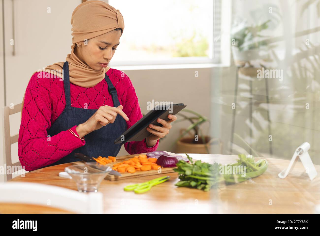 Birassische Frau in Hijab und Schürze, die Gemüse mit Tablette in der Küche zerkleinert Stockfoto