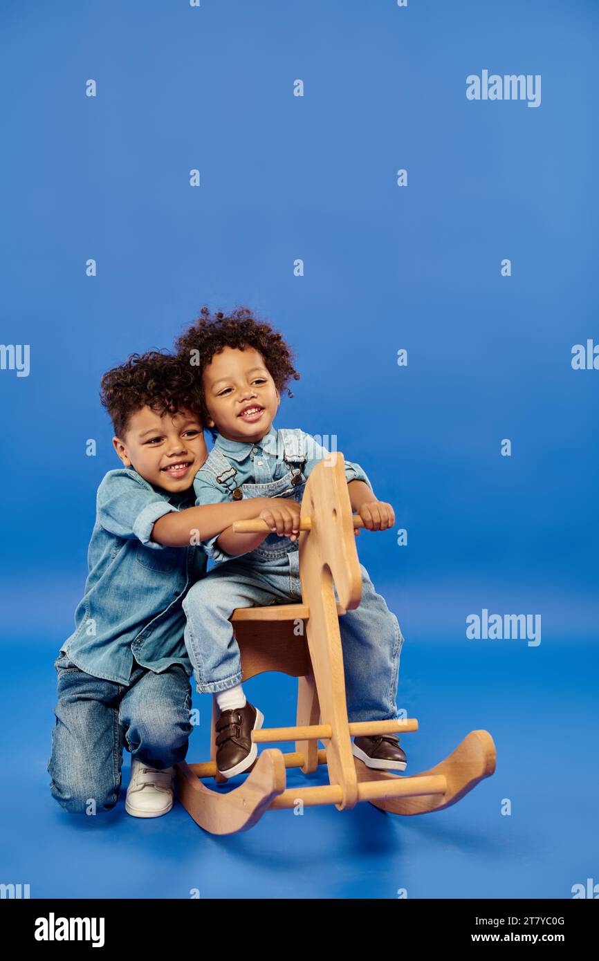 Fröhlicher afroamerikanischer Kleinkind in stylischer Jeanskleidung, der auf einem Schaukelpferd neben Bruder sitzt Stockfoto