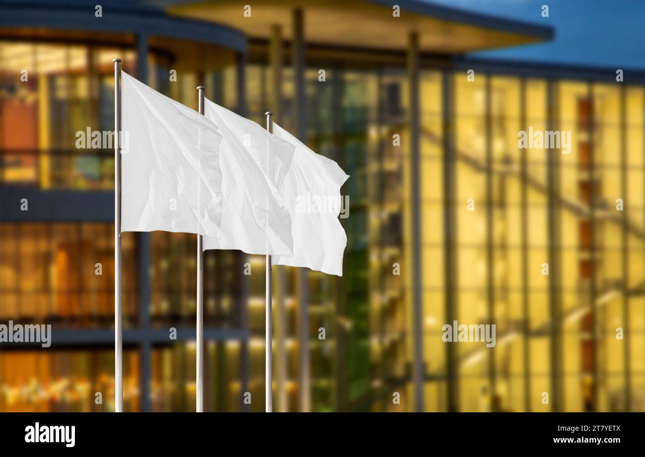 Weiße Firmenflaggen auf Fahnenmasten vor verschwommenem Bürogebäude mit Abendlicht. Leeres Flag-Modell zum Hinzufügen eines Logos, Symbols oder Schildes Stockfoto