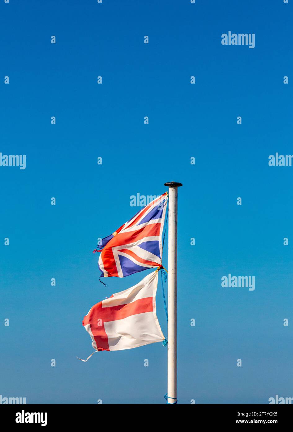 Zerrissene und zerrissene England- und Union Jack-Flaggen auf einem Fahnenmast mit blauem Himmel im Hintergrund. Stockfoto