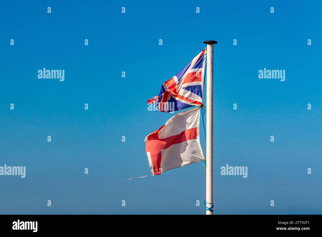 Zerrissene und zerrissene England- und Union Jack-Flaggen auf einem Fahnenmast mit blauem Himmel im Hintergrund. Stockfoto