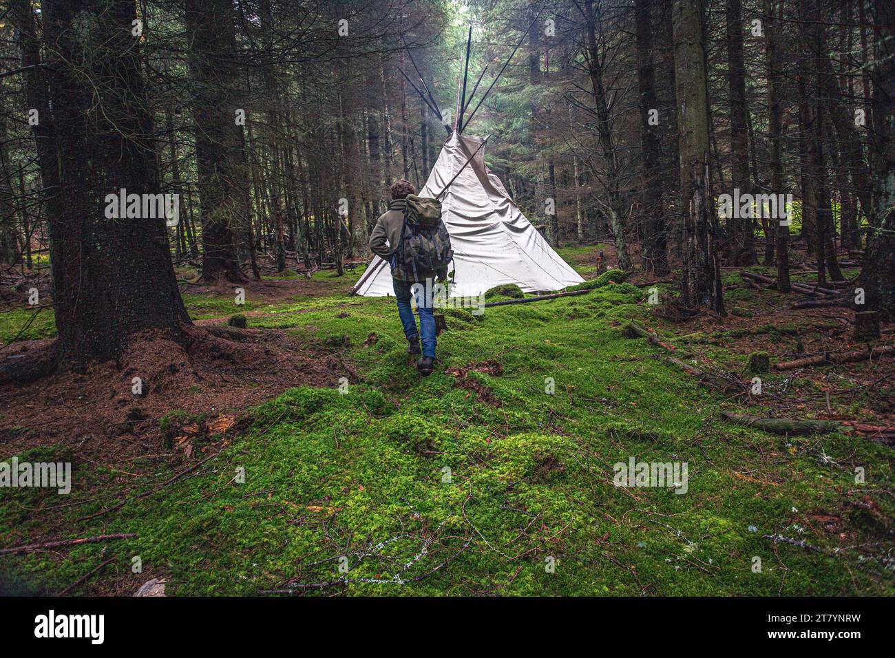 Rückansicht eines Mannes, der mit Rucksack in Richtung Tipi-Zelt in Natur- und Waldlandschaft geht Stockfoto