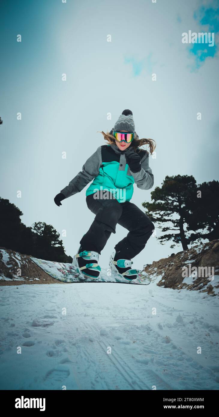 Eine junge Frau mit einer Brille und einem Winteroutfit, die auf einem Snowboard einen schneebedeckten Hügel hinunterreitet Stockfoto