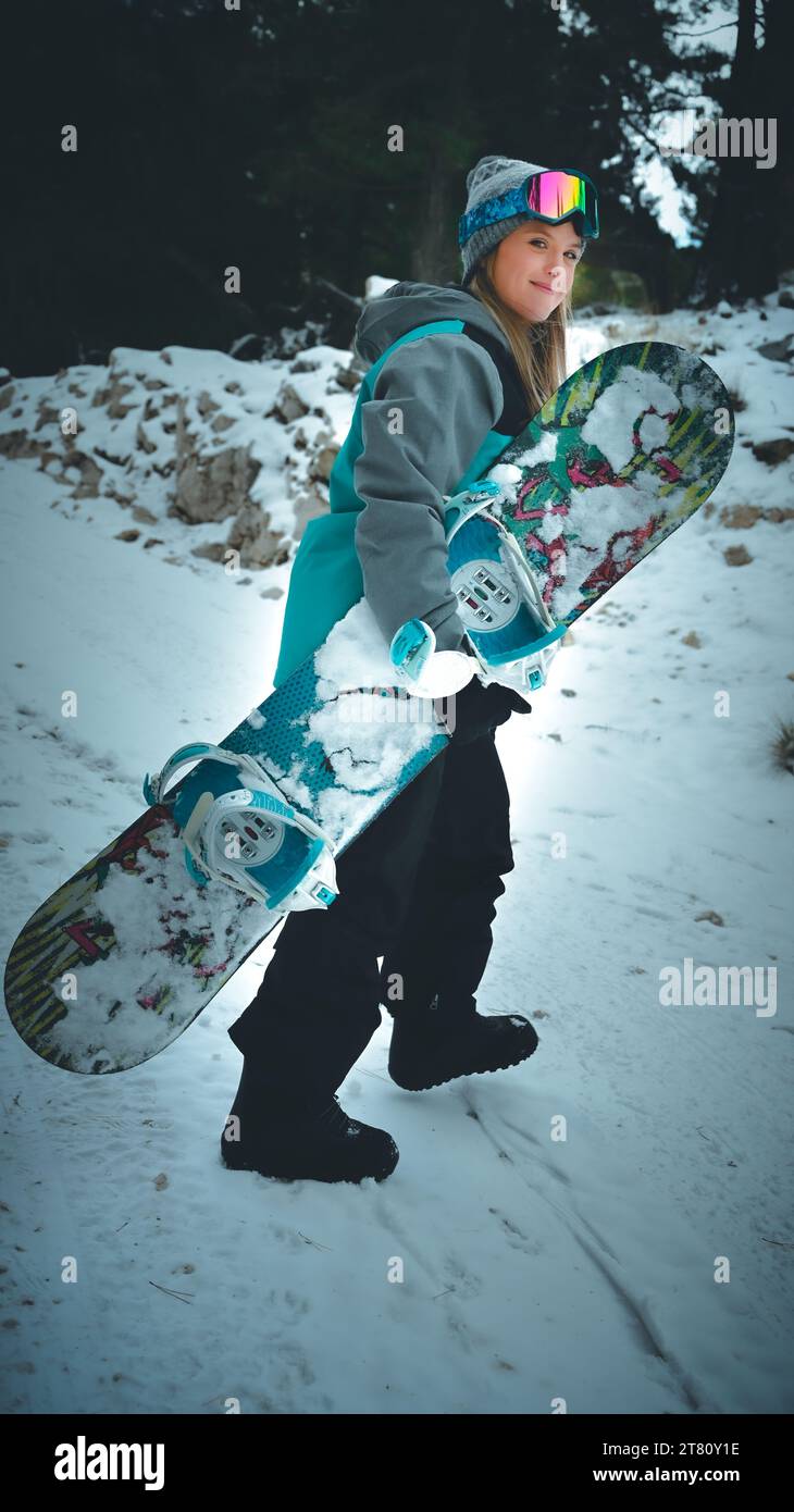 Ein lächelnder junger Mensch steht auf einem schneebedeckten Hügel und hält ein Snowboard in der Hand Stockfoto