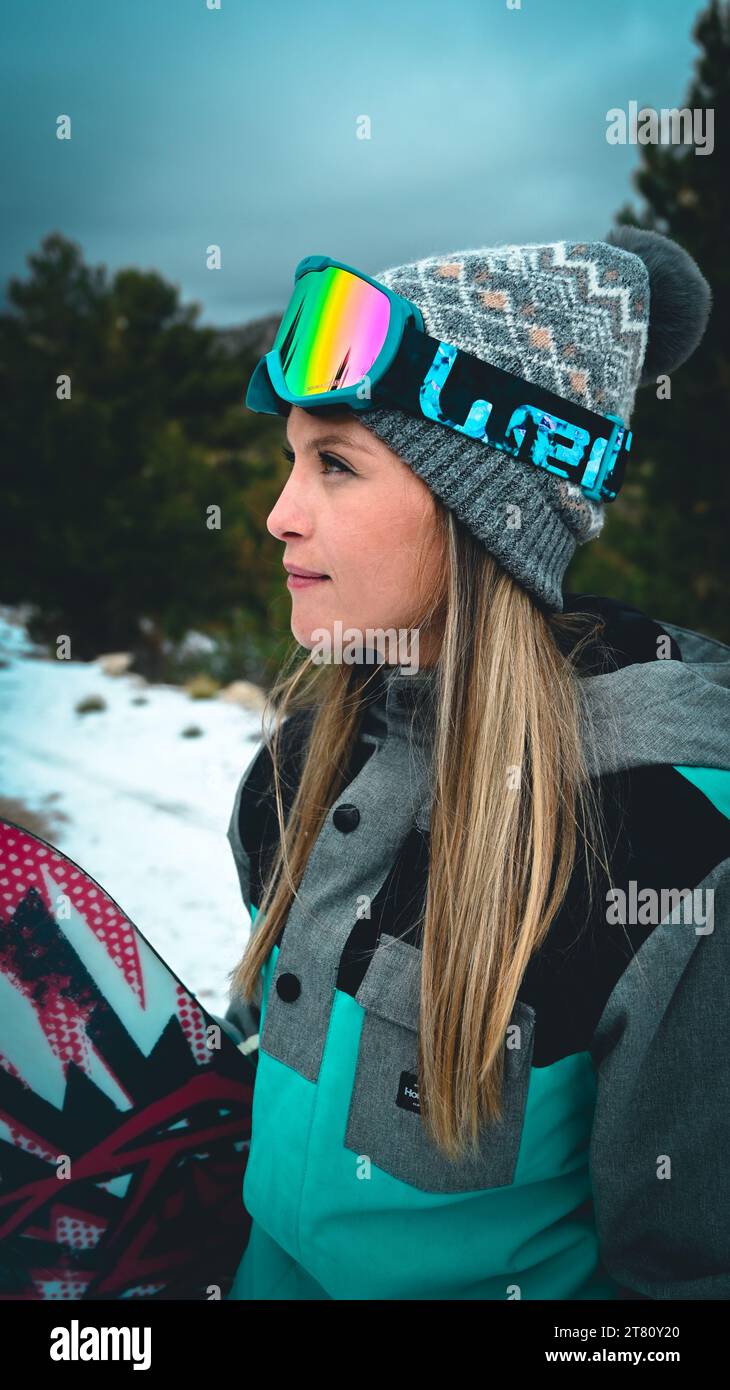 Ein fröhliches Mädchen, das ein Snowboard hält, während es im Schnee steht in einer winterlichen Landschaft, umgeben von immergrünen Bäumen Stockfoto