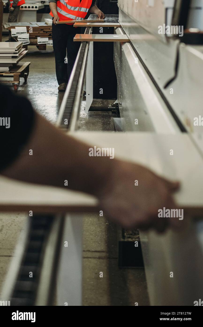 Möbelfabrik. MDF-Holzteile, die sich entlang eines speziell ausgerüsteten Förderbandes bewegen. PVC-Kanten. Ein Mann legt mit den Händen einen Möbelteller auf die Maschine Stockfoto
