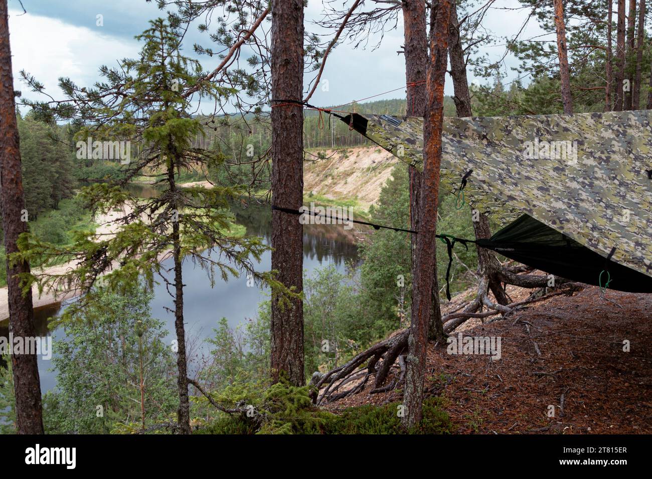 Auf einem Wanderweg im Norden Finnlands befindet sich eine Hängematte mit Plane zwischen den Bäumen mit Blick auf einen Fluss unterhalb des Waldes Stockfoto