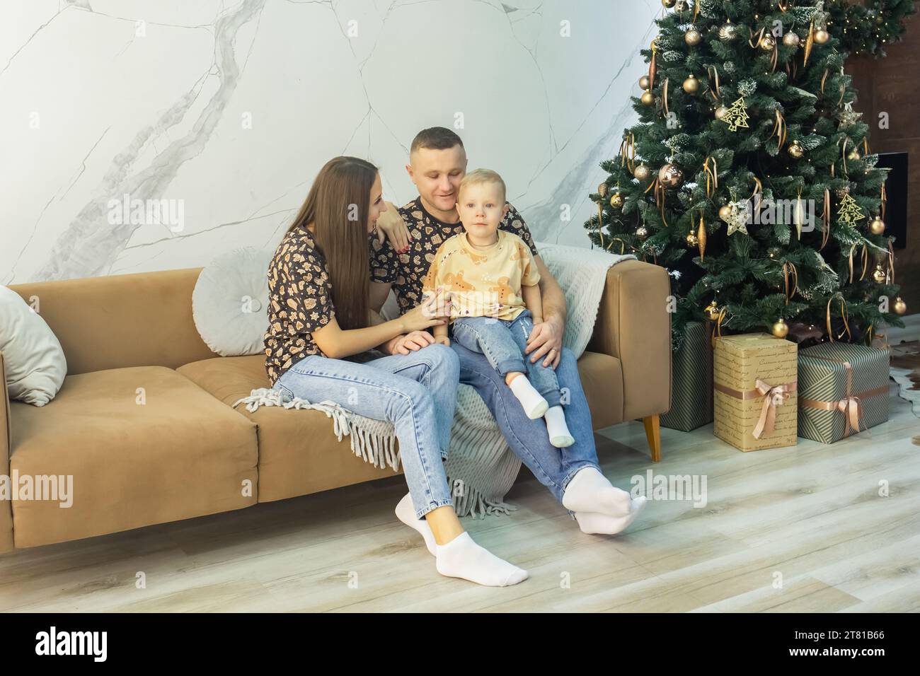 Wunderschöne Familie, die auf dem Sofa im Wohnzimmer sitzt, dekoriert für Weihnachten und Neujahr. Junge Familie in der Nähe eines schönen und großen Neujahrsbaums Stockfoto