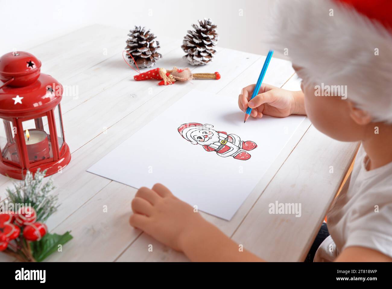 Ein junges Mädchen, das den Weihnachtsmann auf Papier mit festlichem Hut zeichnet. Weihnachtsdekoration auf weißem Holztisch. Kreatives Urlaubskonzept Stockfoto