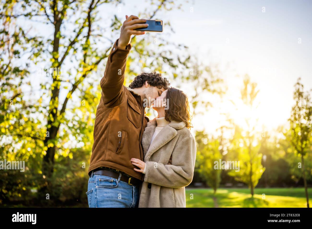 Porträt eines glücklichen Liebespaares im Park bei Sonnenuntergang. Sie küssen und machen Selfie. Stockfoto