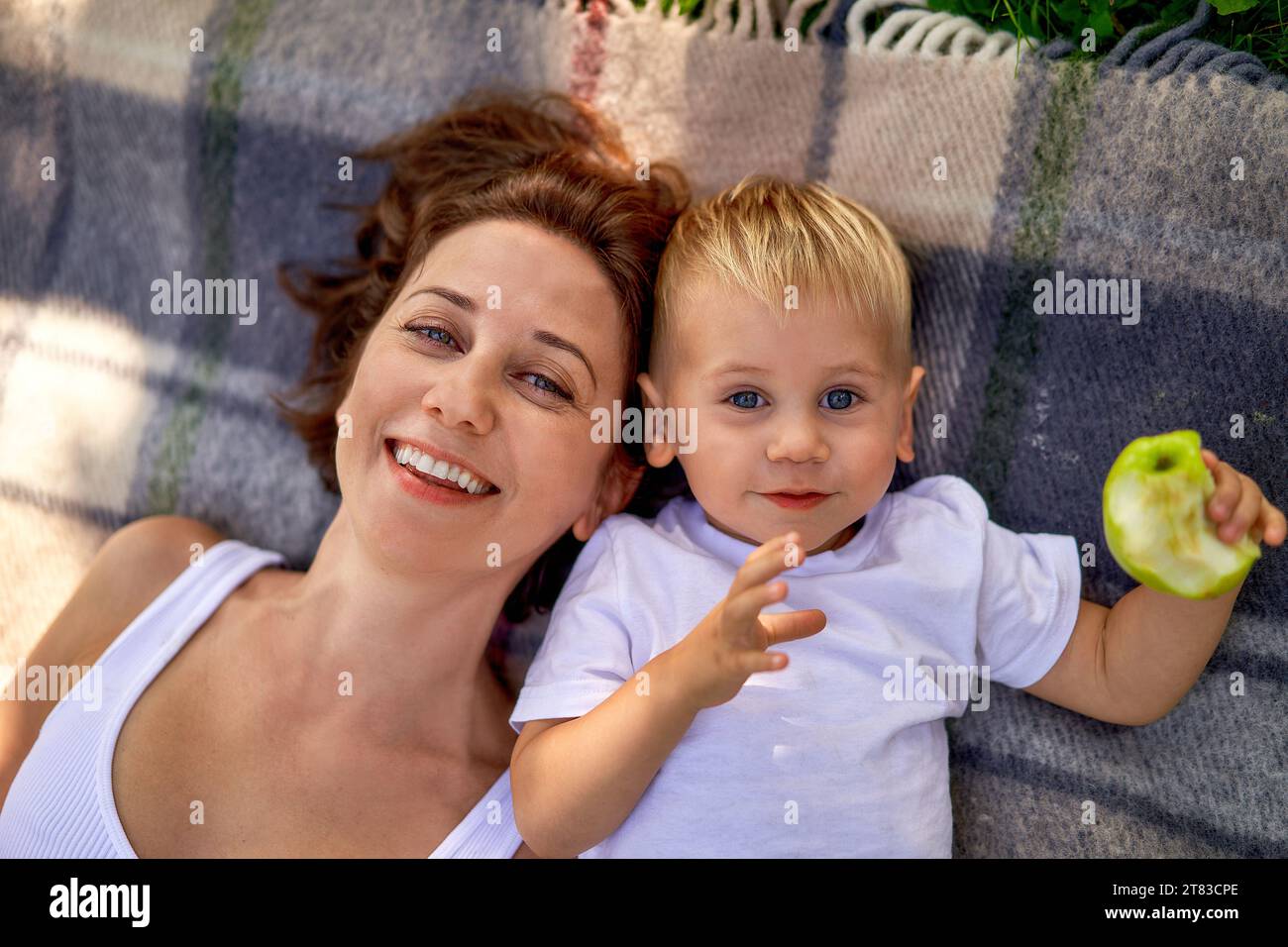 Eine junge europäische Mutter mit ihrem niedlichen kleinen Sohn, der auf dem Bett liegt, kariert und in die Kamera lächelt, von oben. Ein Kind hat einen gebissenen grünen Apfel in der Hand. Das Konzept der Erziehung und des gemeinsamen Zeitverhaltens in der Natur. Das Glück der Mutterschaft, Blick von oben Stockfoto