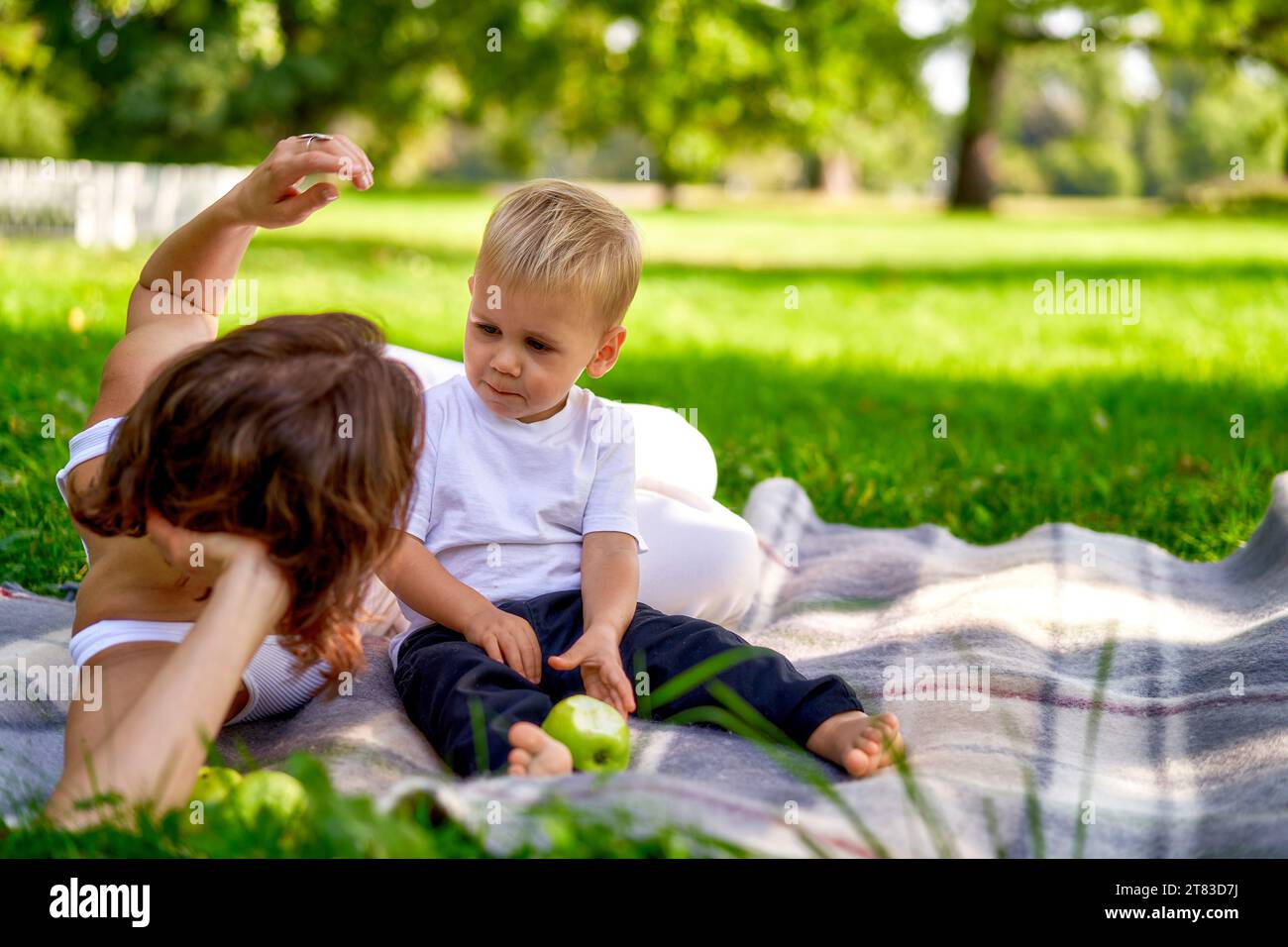 Mama in weißen Kleidern ruht mit ihrem Kind im Park auf grünem Gras. Gute Eltern draußen. Lifestyle. Stockfoto
