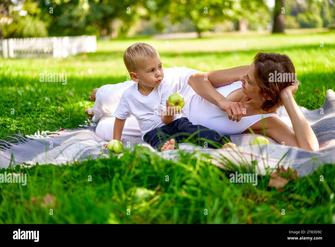 Mama in weißen Kleidern ruht mit ihrem Kind im Park auf grünem Gras. Gute Eltern draußen. Lifestyle. Stockfoto