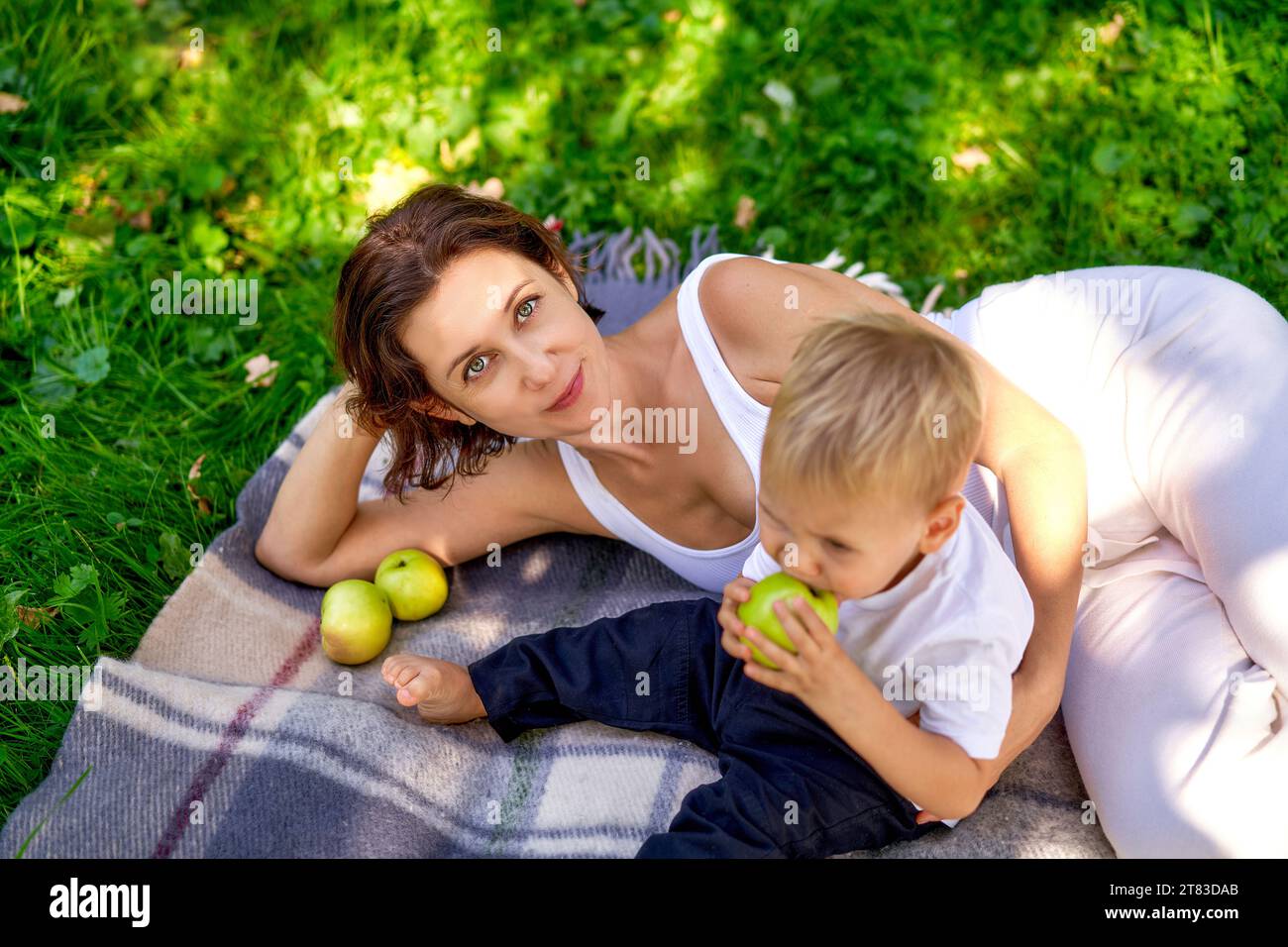 Eine junge europäische Mutter mit ihrem niedlichen kleinen Sohn, der auf dem Bett liegt, kariert und in die Kamera lächelt. Ein Kind hat einen gebissenen grünen Apfel in der Hand. Das Konzept der Erziehung und des gemeinsamen Zeitverhaltens in der Natur. Das Glück der Mutterschaft, Blick von oben Stockfoto