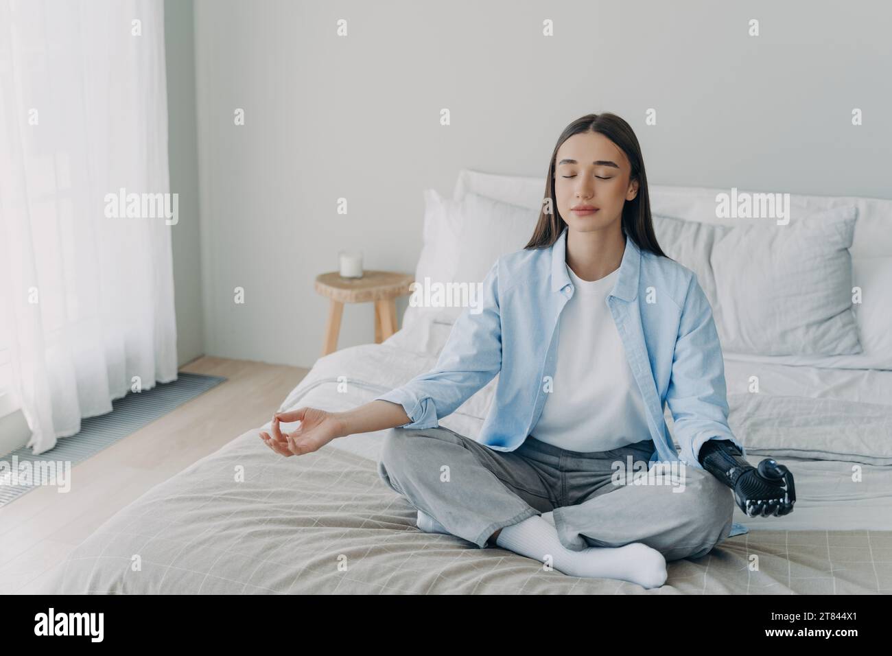 Ruhige junge Frau mit prothetischer Hand, die auf dem Bett meditiert, Frieden und Belastbarkeit verkörpert Stockfoto