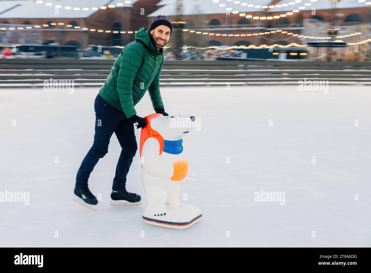 Fröhlicher Mann mit Eisbären-Eislaufhilfe auf einer Eisbahn mit festlichen Lichtern Stockfoto