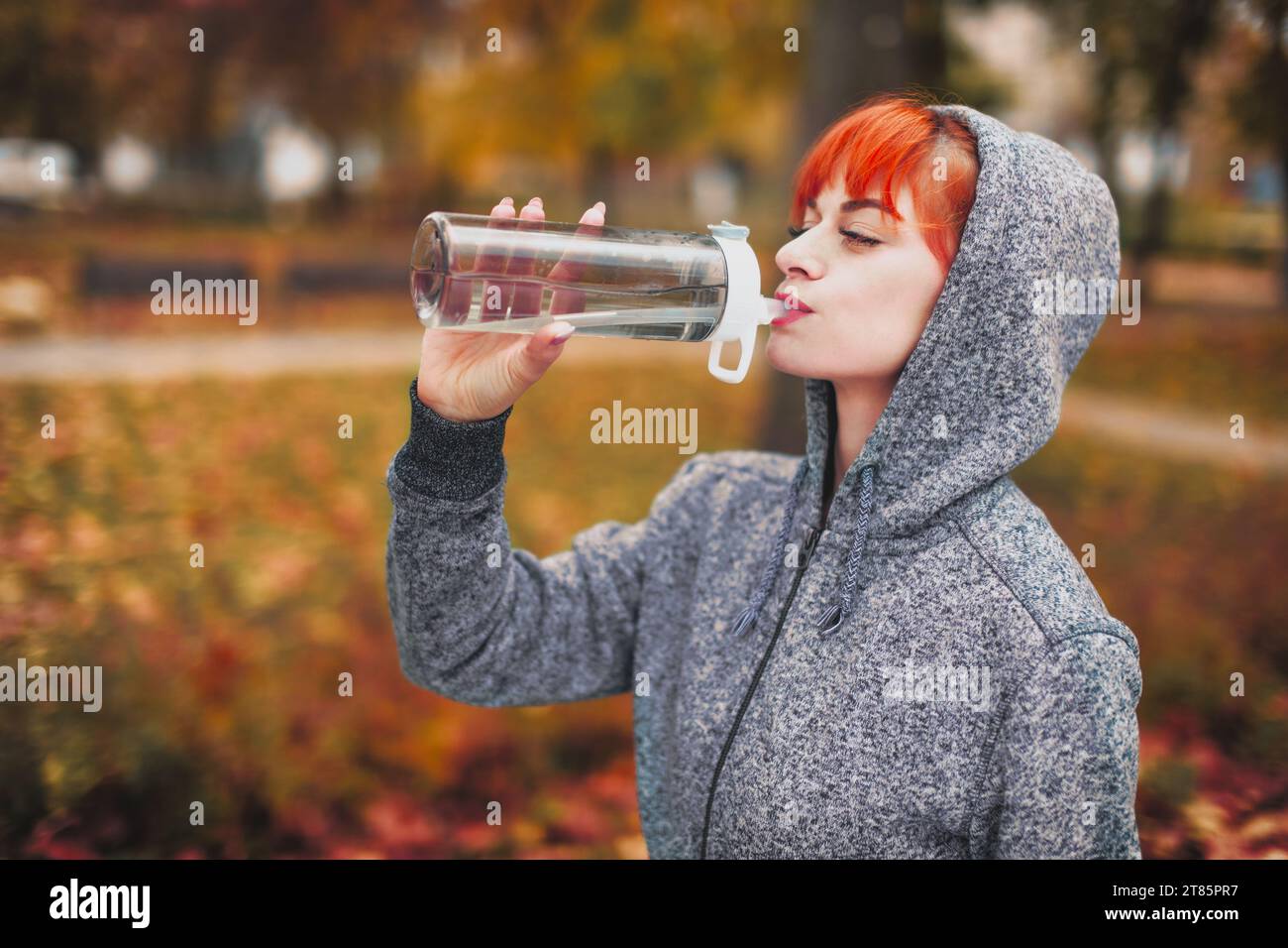 Junge Rothaarige Athletin Frau im Kapuzenpullover Trinkwasser aus der Flasche während der Outdoor-Vorbereitung im Park Stockfoto