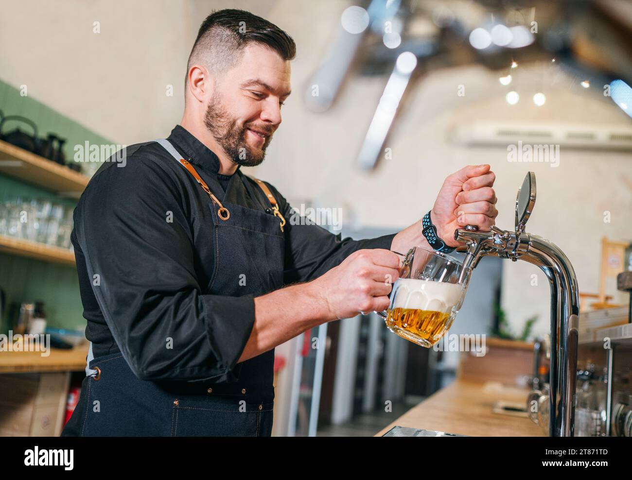 Ein lächelnder, stilvoller Bärenbarmann in schwarzer Uniform mit einer Schürze, der frisches Lagerbier in einen Glasbecher an der Bar zapft. Erfolgreiche Leute, Bierkonzern Stockfoto