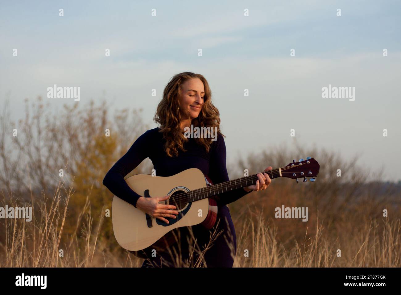 Schöne junge, lächelnde Frau, die eine Akustikgitarre spielt, während sie im Feld zwischen hohem, getrocknetem Gras steht. Talentierter Musiker. Sonnenuntergang im Herbst. Romantische Mo Stockfoto