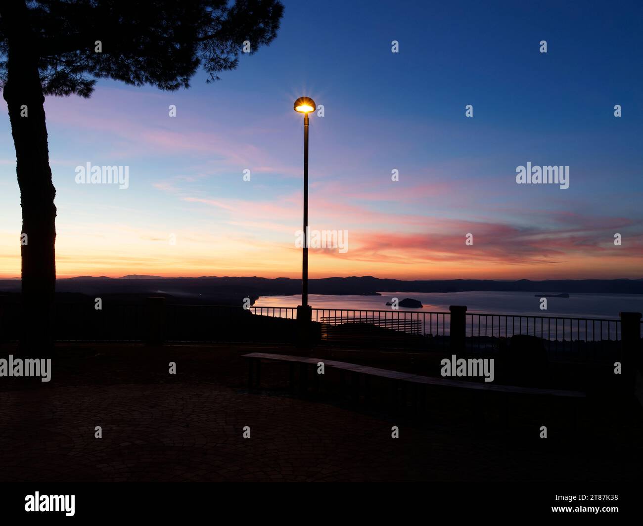 Blick auf den ruhigen See Bolsena, während die Sonne von einem Aussichtspunkt in Montefiascone mit einer und Straßenlaterne untergeht. Region Latium, Italien. November 2023 Stockfoto