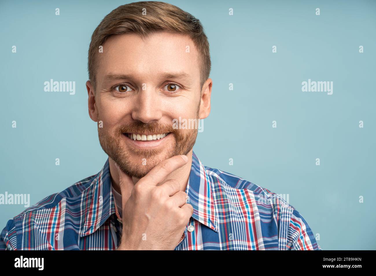 Porträt kaukasischer bärtiger Mann, der in die Kamera schaut, lächelnd, berührendes Kinn auf blauem Hintergrund. Stockfoto