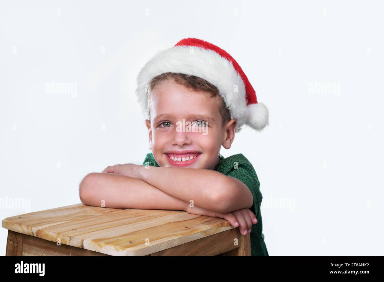Der fünfjährige kaukasische Junge mit einem fröhlichen Lächeln im Gesicht, der in die Kamera schaut, liegt mit gefalteten Armen auf einem Holztisch Stockfoto