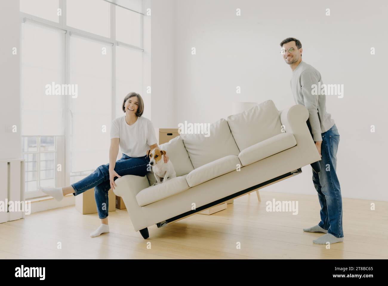 Ein glückliches Paar mit ihrem Hund, der ein Sofa in einem großen, sonnendurchfluteten Raum mit hohen Fenstern bewegt Stockfoto