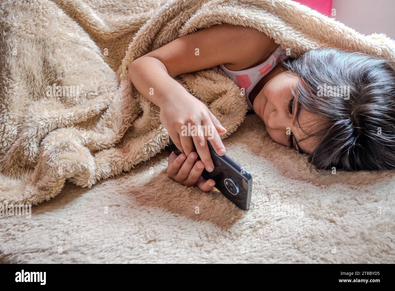 Süßes kleines Mädchen, das ein Smartphone benutzt. Junge, der mit dem Handy spielt, auf einem Bett liegt. Freizeit. Technologie und Internetkonzept. Kleinkind liegt auf Bett A Stockfoto