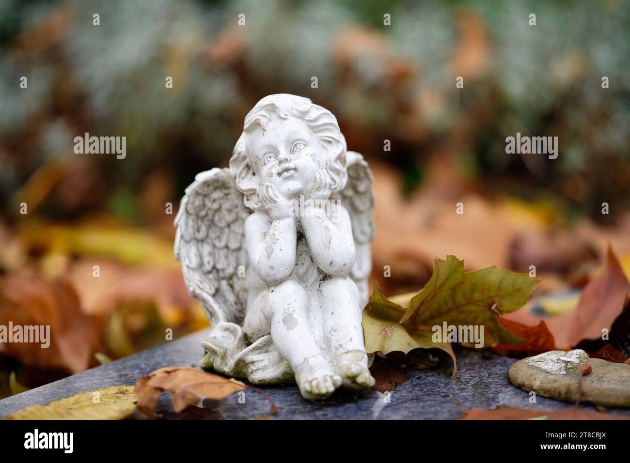 Eine kleine weiße Engelsfigur sitzt träumend auf einem Grabstein mit Herbstlaub Stockfoto