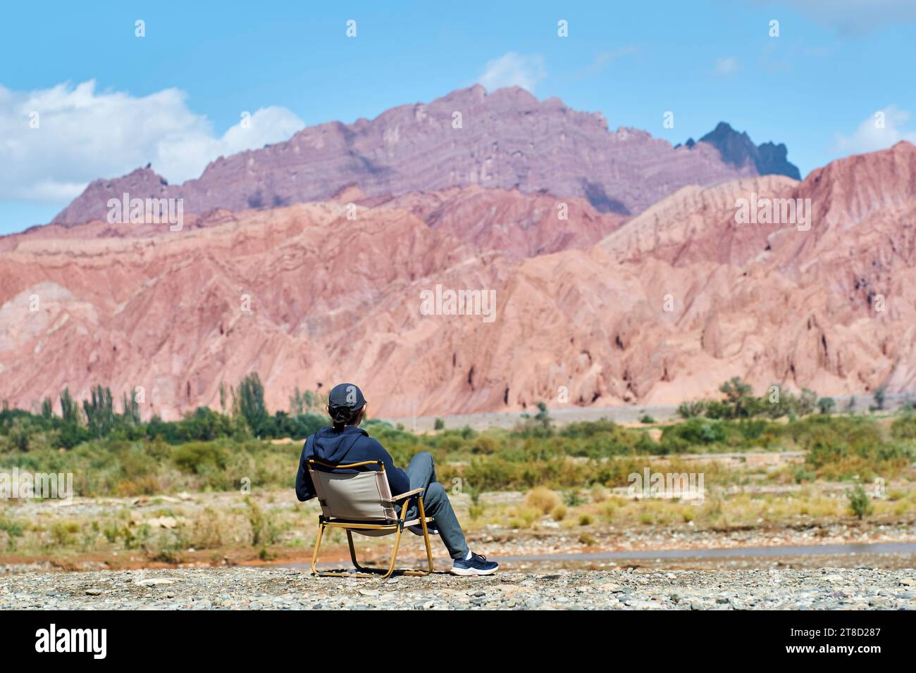 Rückansicht eines asiatischen Mannes, der im Stuhl auf dem Flussbett sitzt und auf die Klippen der Schlucht in Xinjiang, China, blickt Stockfoto