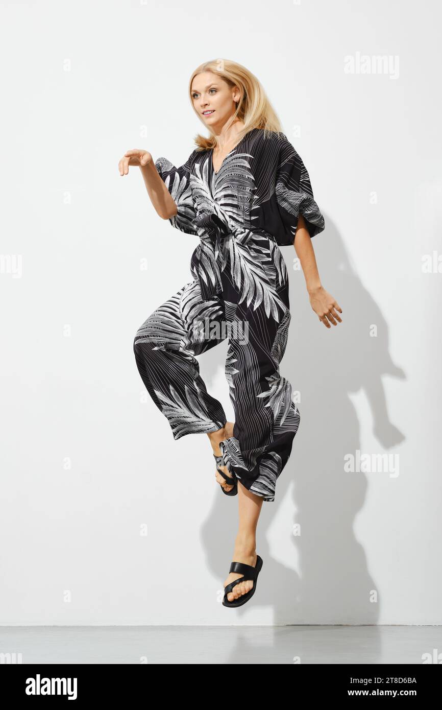 Verspielte blonde Frau in gemustertem Overall, die drinnen springt Stockfoto