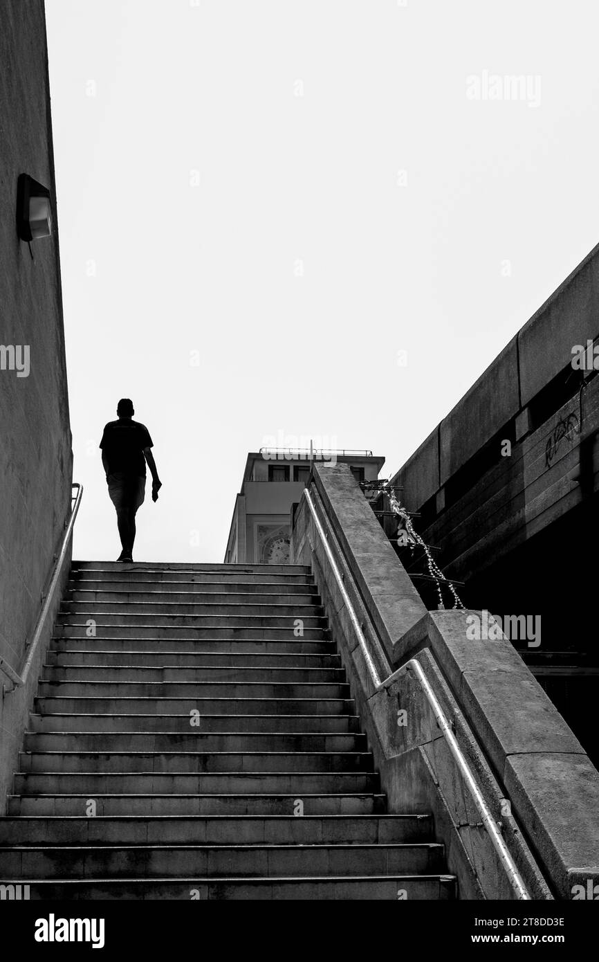 Schwarz-weißes Bild der Silhouette eines Mannes, der draußen eine breite Betontreppe vor hellem Himmel hinaufgeht. Stockfoto