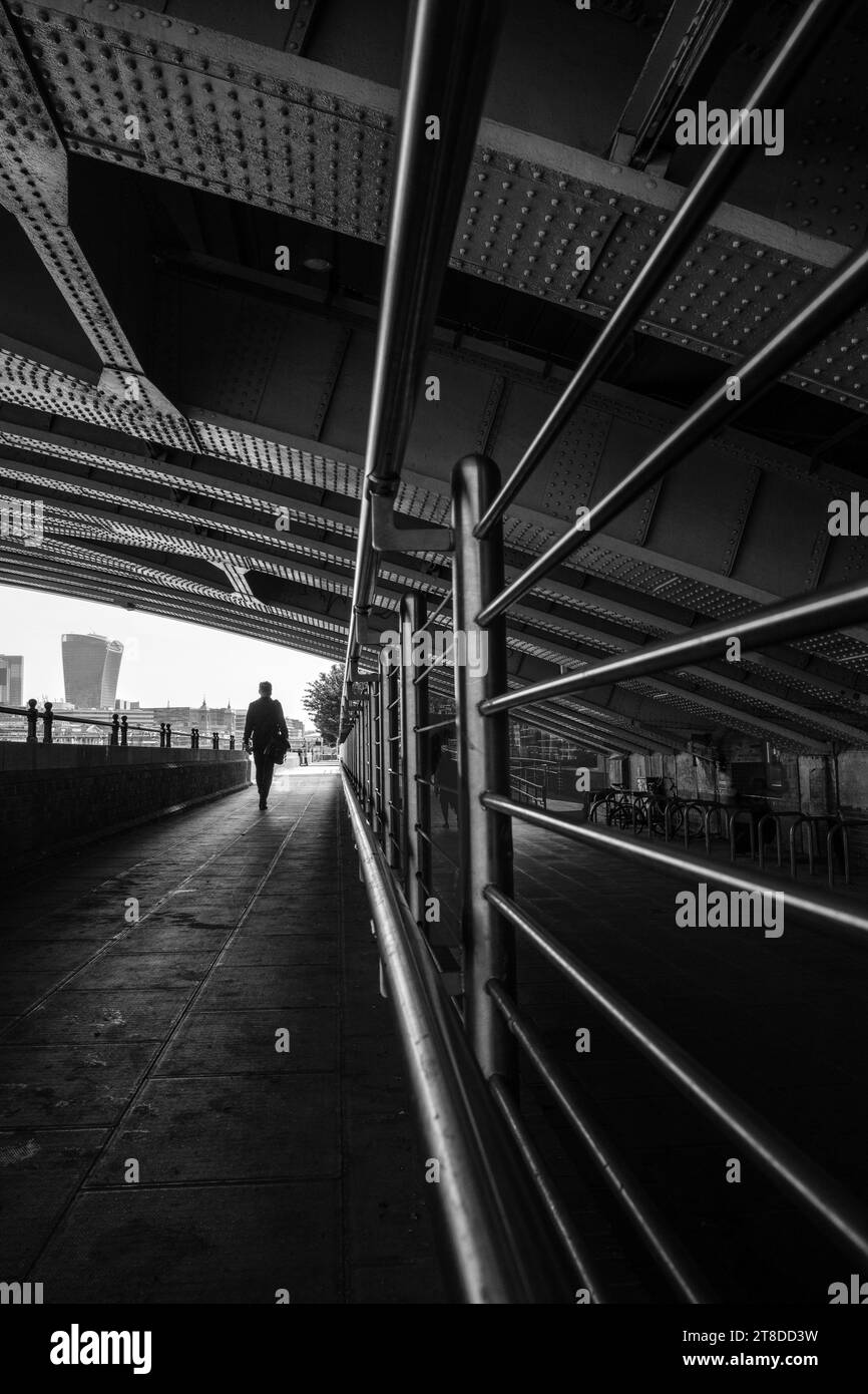 Schwarzweißbild. Silhouette eines Mannes, der durch einen Durchgang mit niedrigem Dach und führenden Linien geht, vor hellem Himmel mit Walkie-Talkie-Gebäude. Stockfoto