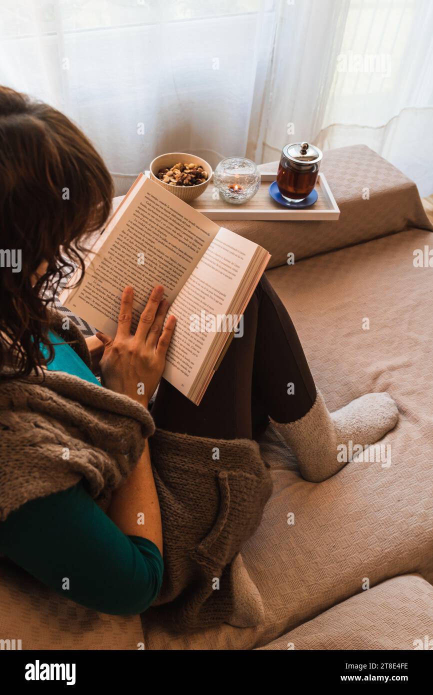 Vertikales Foto, eine Erwachsene Frau, kaukasisch, lässige Kleidung, sitzt auf dem Sofa, neben dem Fenster, mit einem offenen Buch in der Hand, mit einem Tee. Stockfoto
