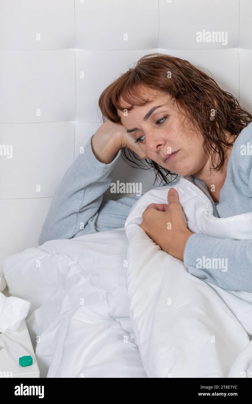 Vertikales Foto, mittelErwachsene kaukasische Frau, mit langem blauen Pyjama, krank im Bett, bedeckt mit einer weißen Decke. Stockfoto