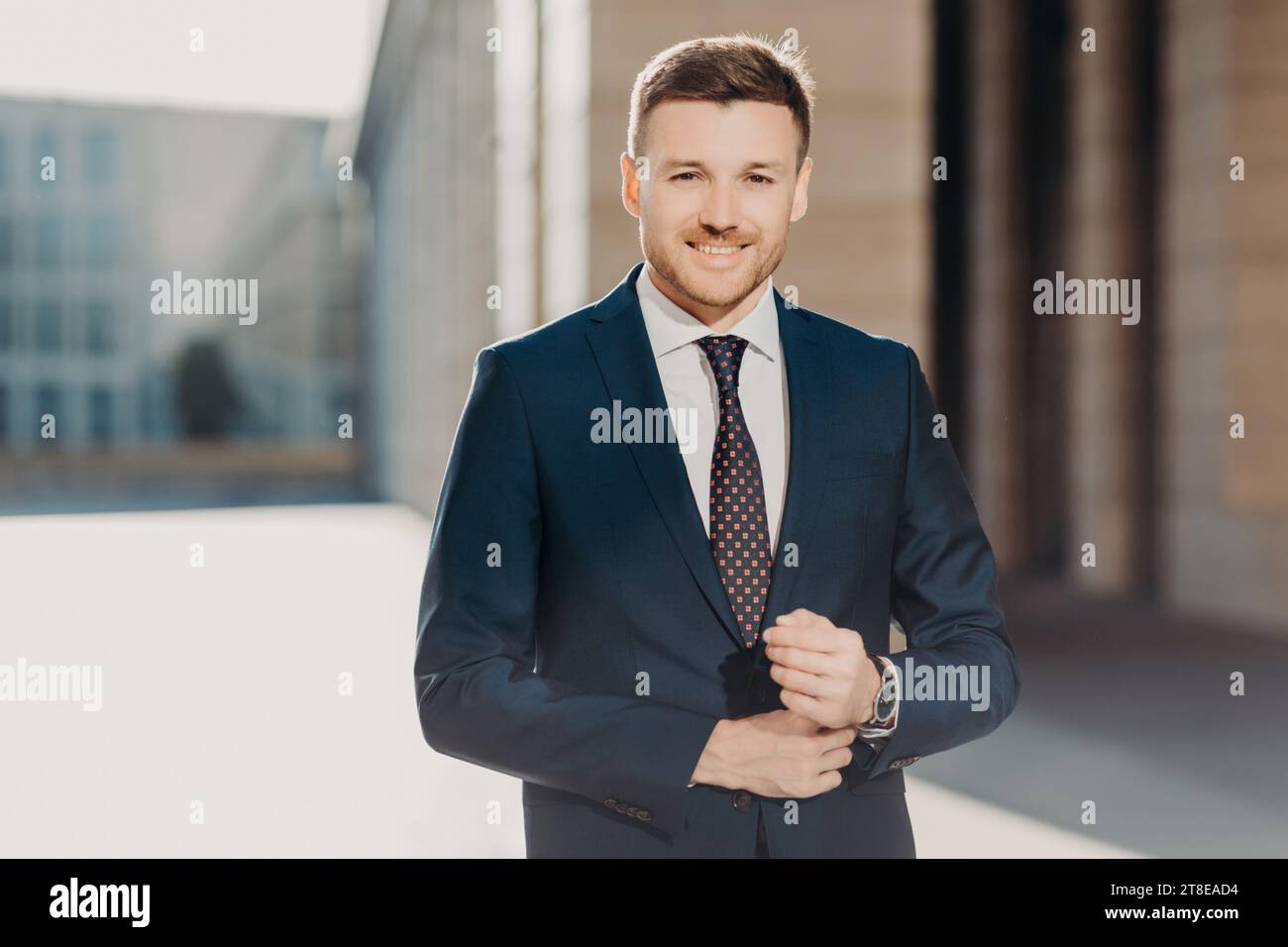 Selbstbewusster Geschäftsmann, der seine Anzugsjacke im Freien mit einer modernen Gebäudefassade im Hintergrund zuknöpft Stockfoto