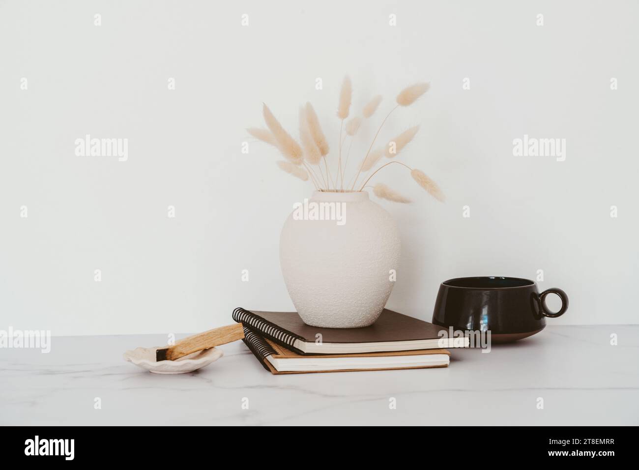 Stilleben von trockenen Lagurus-Blüten in Vase, Tasse Tee, Notizbücher und palo santo auf weißem Tisch. Kopierbereich. Vorderansicht Stockfoto