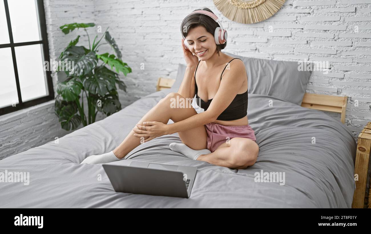Entspannte, wunderschöne junge hispanische Frau, die Musik hört, während sie sich auf ihrem Bett entspannt und in ihrem gemütlichen Schlafzimmer in Morgenlicht getaucht ist Stockfoto Entspannte, wunderschöne junge hispanische Frau, die Musik hört, während sie sich auf ihrem Bett entspannt und in ihrem gemütlichen Schlafzimmer in Morgenlicht getaucht ist Stockfoto