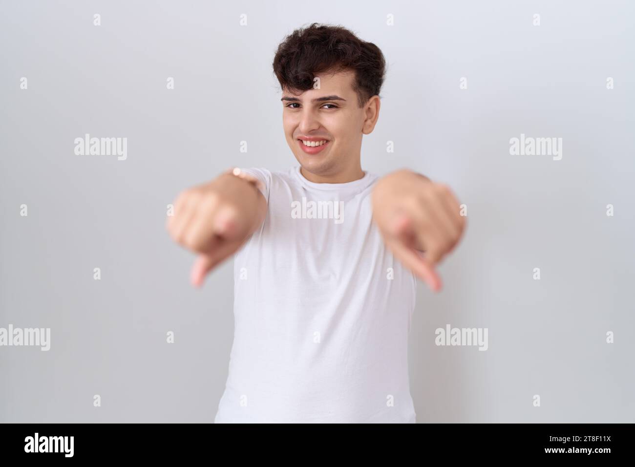 Junger nicht binärer Mann, der ein lässiges weißes T-Shirt trägt, das mit den Fingern auf dich und die Kamera zeigt, positiv und fröhlich lächelnd Stockfoto