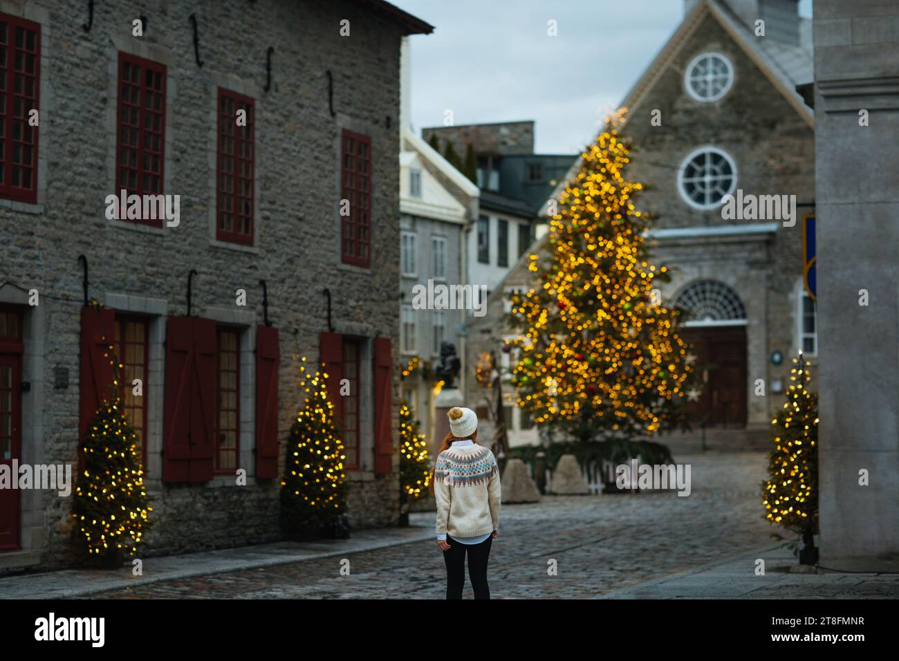 Die Rückansicht der Frau mit warmem Hut und Pullover zeigt die funkelnde Weihnachtsdekoration und einen großen Weihnachtsbaum auf einem Kopfsteinpflaster Stockfoto