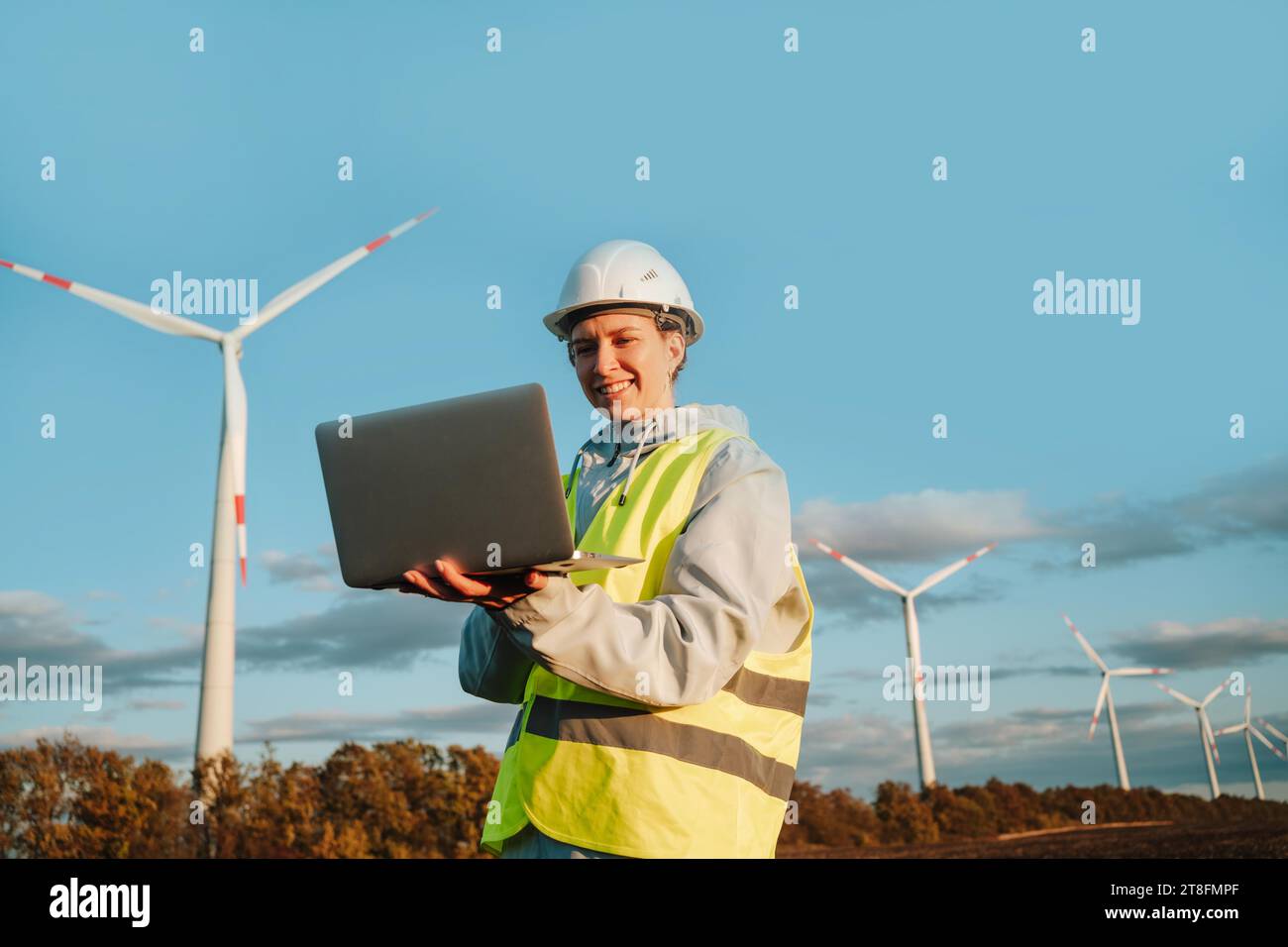 Eine Ingenieurin in Sicherheitsausrüstung verwendet einen Laptop mit Windturbinen im Hintergrund, die erneuerbare Energien und Technologie repräsentieren. Stockfoto