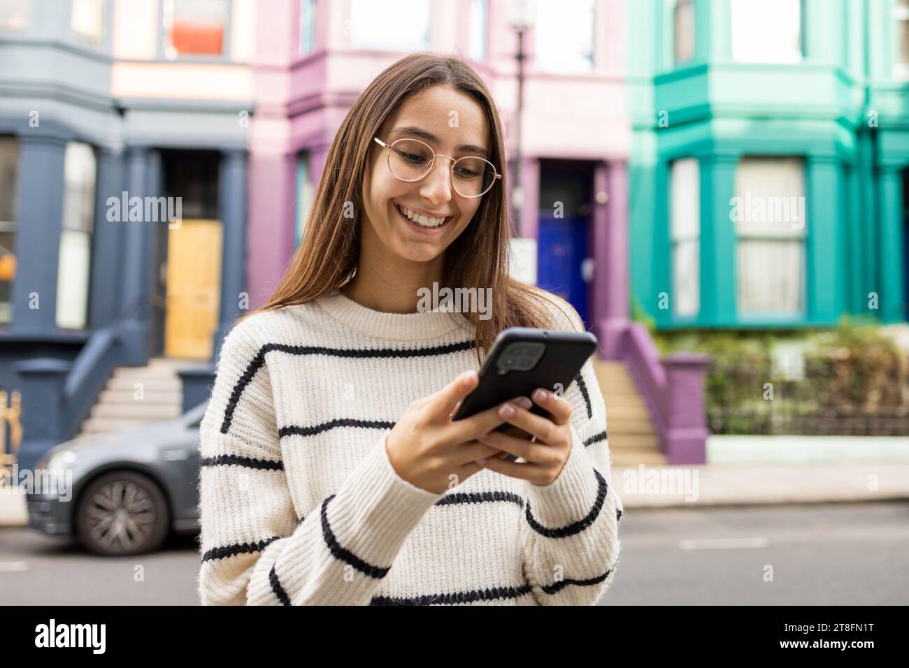 Fröhliche junge Frau in lässiger Kleidung genießt ihr Smartphone vor einer lebendigen, farbigen Kulisse in London. Stockfoto