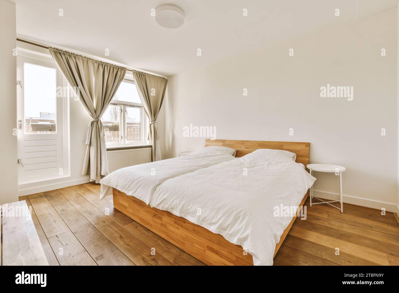 Komfortables Bett mit Hocker vor hellem Fenster in luxuriösem Schlafzimmer in einem modernen Apartment mit weißen Wänden Stockfoto