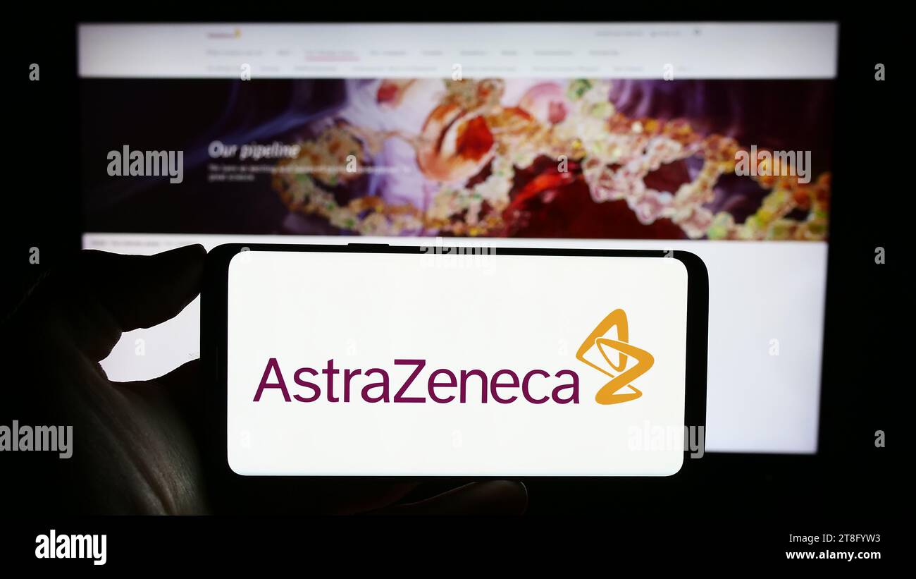 Person, die ein Mobiltelefon mit dem Logo des Pharmaunternehmens AstraZeneca plc vor der Business-Webseite hält. Schwerpunkt auf dem Display des Telefons. Stockfoto