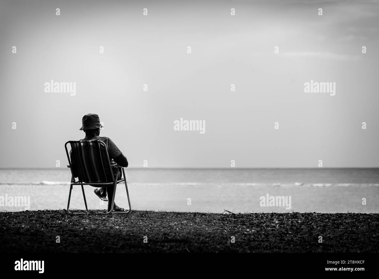 Künstlerisches Schwarz-weiß-Bild eines einsamen Mannes, der auf einem Campingstuhl am Strand sitzt und ein Buch liest. Stockfoto