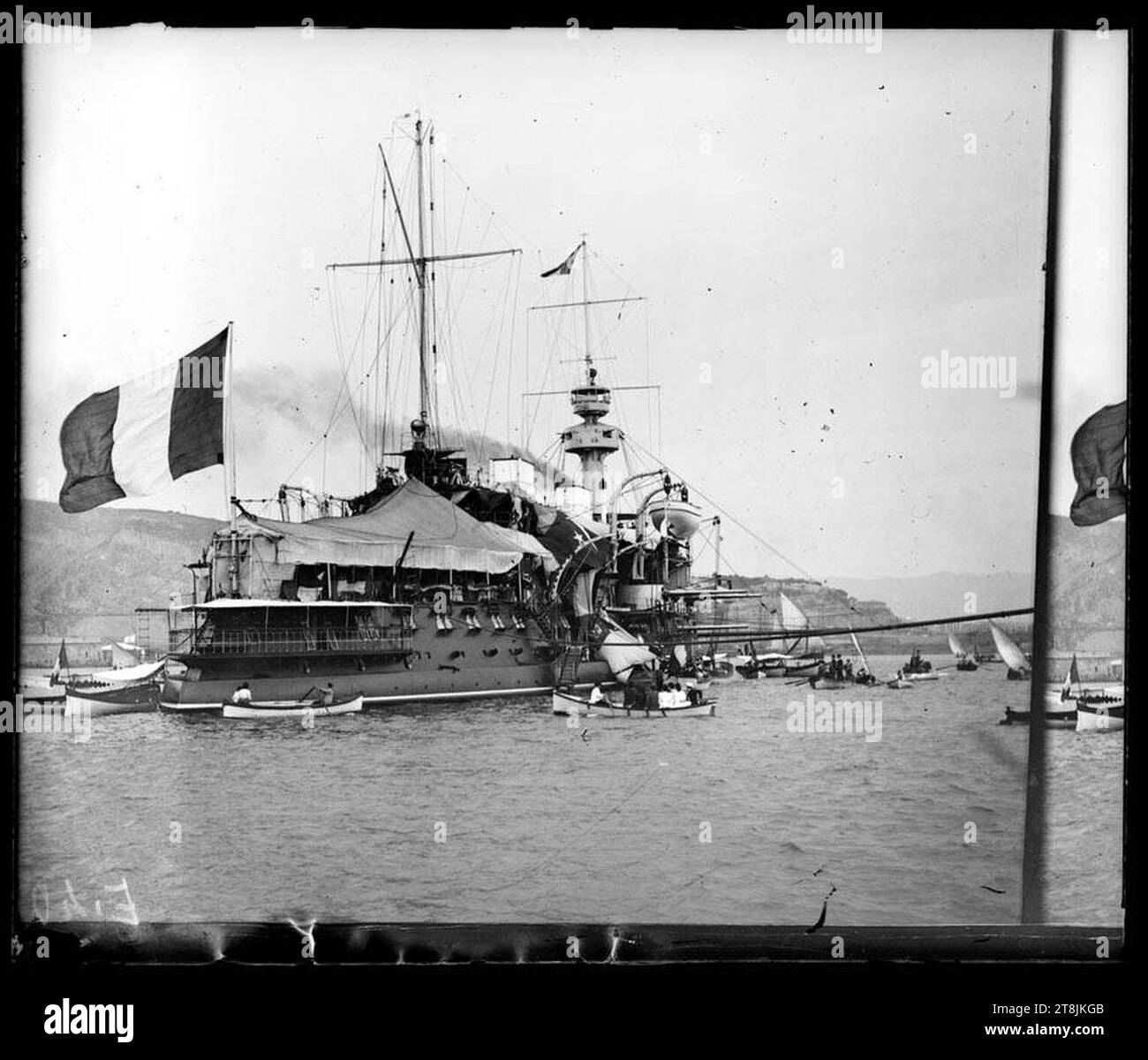 Vaixell amb bandera francesa o italiana voltat de barques, amb un Port i una muntanya al fons Stockfoto