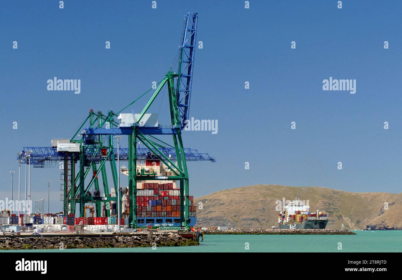 Port Lyttleton. NZ, Containerkrane bestehen aus einem Traggerüst, das die Länge eines Kais oder Werks auf einer Schiene überqueren kann. Anstelle eines Hakens sind sie mit einem speziellen Handlingwerkzeug ausgestattet, das als Spreuverteiler bezeichnet wird. Der Spreuverteiler kann auf einen Behälter abgesenkt werden und verriegelt sich mit einem Twistlock-Mechanismus an den vier Verriegelungspunkten des Behälters („Eckgussteile“). Kräne transportieren normalerweise einen einzelnen Container auf einmal, aber einige neuere Kräne können zwei bis vier 20-Fuß-Container auf einmal aufnehmen Stockfoto