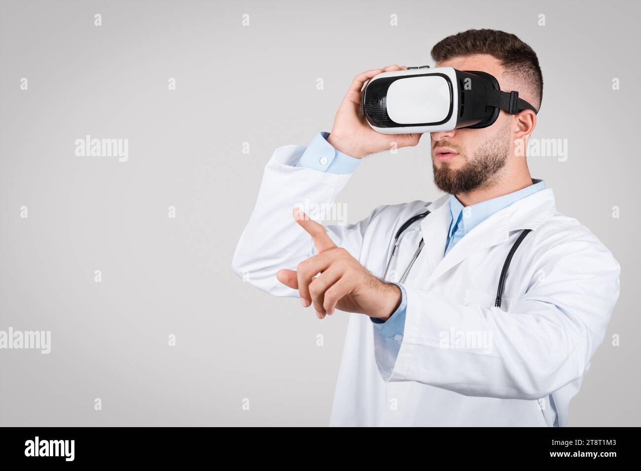Junger männlicher Arzt im weißen Mantel mit VR-Brille, grauer Hintergrund Stockfoto