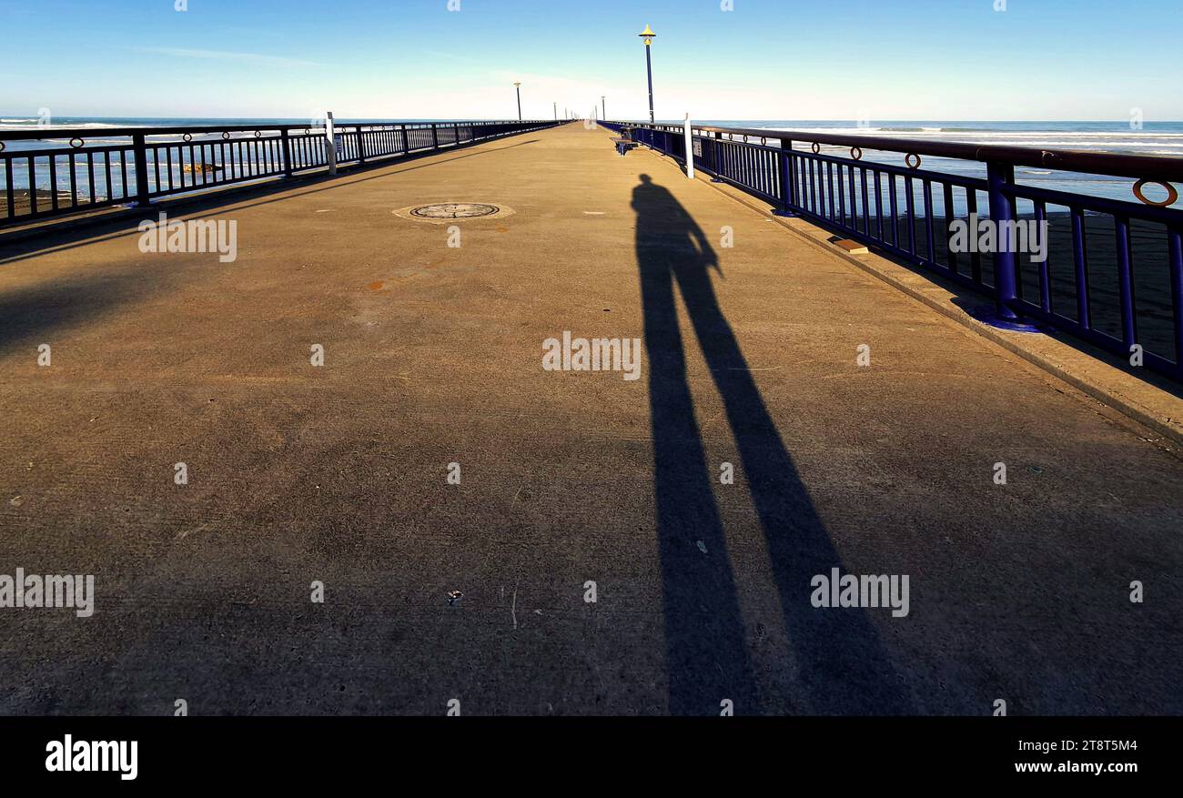 In den Schatten gelangten zwei New Brighton Piers in New Brighton, Neuseeland. Der erste Pier in Holzbauweise wurde am 18. Januar 1894 eröffnet und am 12. Oktober 1965 abgerissen. Der heutige Betonpfeiler wurde am 1. November 1997 eröffnet. Es ist eine der Ikonen von Christchurch, Neuseeland Stockfoto