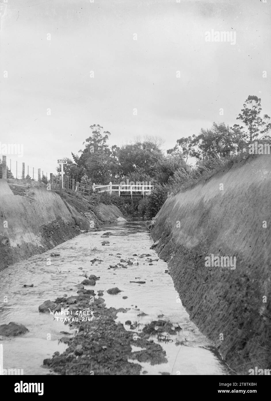 Waipipi Creek, Tuakau, Aussicht auf den Bach hinunter zu einer Straßenbrücke in der Ferne. Es gibt ein "Stop"-Schild am linken Damm, möglicherweise zwischen 1910-1925 Stockfoto